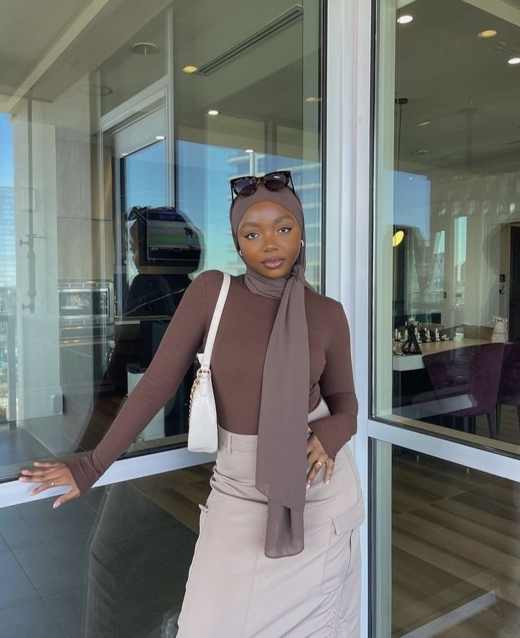 Rodia in a brown top and taupe hijab, leaning on a glass door with sunglasses on her head.