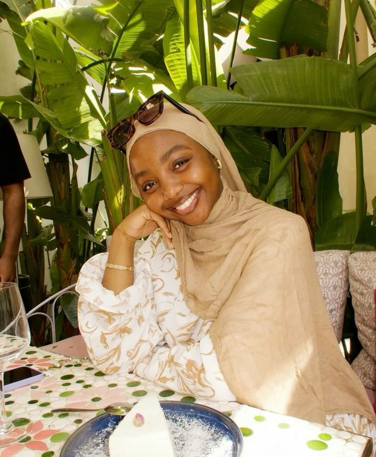 Rodia smiling at a café table surrounded by greenery, in a soft beige hijab.