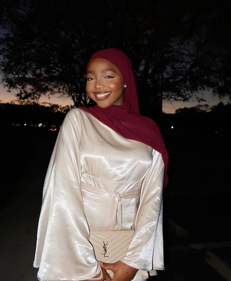 Rodia smiling outdoors at dusk in a satin cream outfit and deep red hijab.
