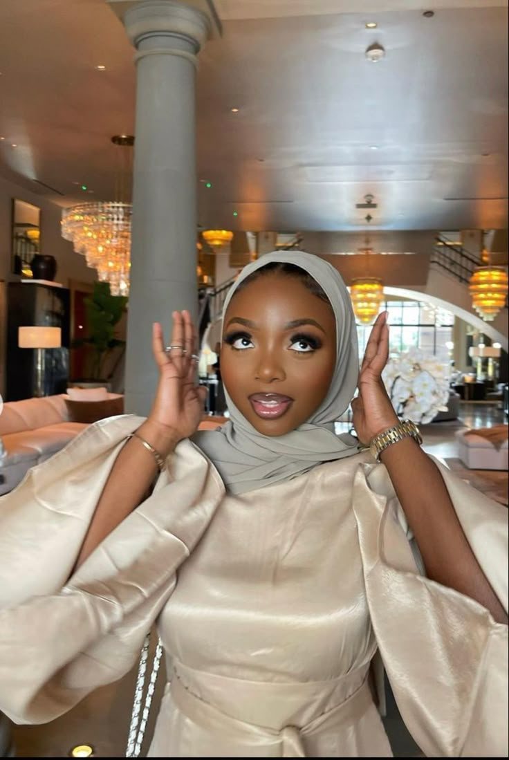 Rodia framing her face with both hands, in a champagne outfit and sage hijab, in an elegant hotel lobby.