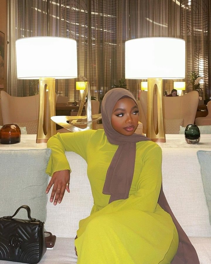 Rodia seated on a sofa between two lamps, in a bright green dress and brown hijab.