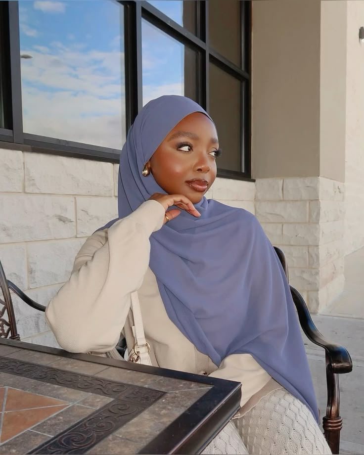 Rodia seated by a window in a lilac hijab, chin resting on her hand, looking to the side.