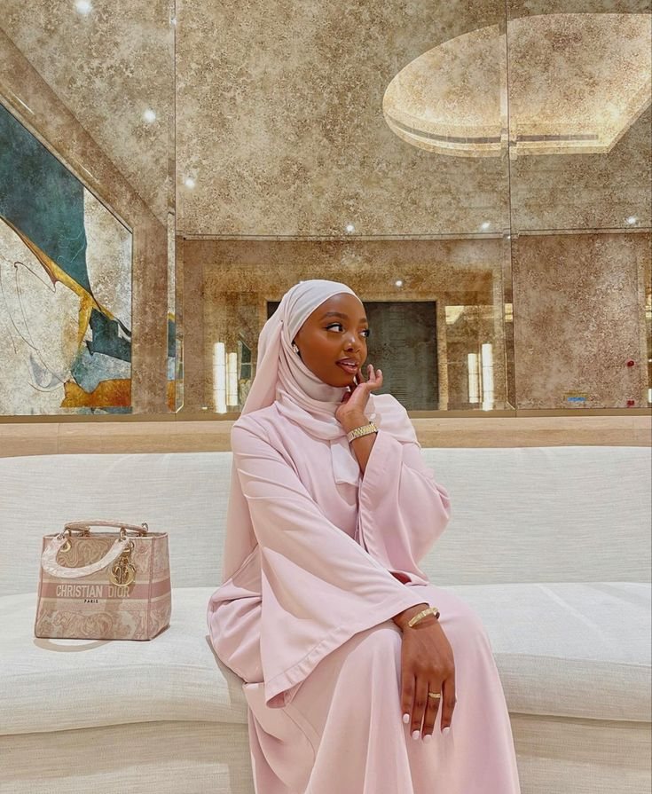 Rodia in a pale pink abaya and hijab, seated on a white sofa with a quilted bag beside her.